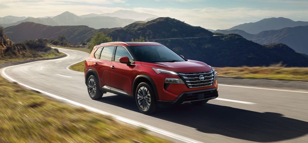 A red 2025 Nissan Rogue driving on a winding road through a mountainous landscape under a hazy blue sky.