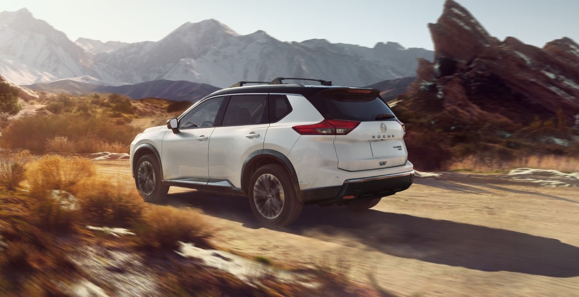 A white 2025 Nissan Rogue driving on a dusty road with mountains and rocky terrain in the background.