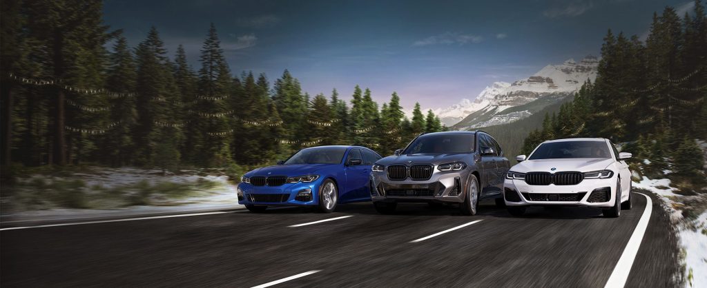 Three BMW vehicles driving on a mountain road surrounded by pine trees and snow-covered peaks in the background.